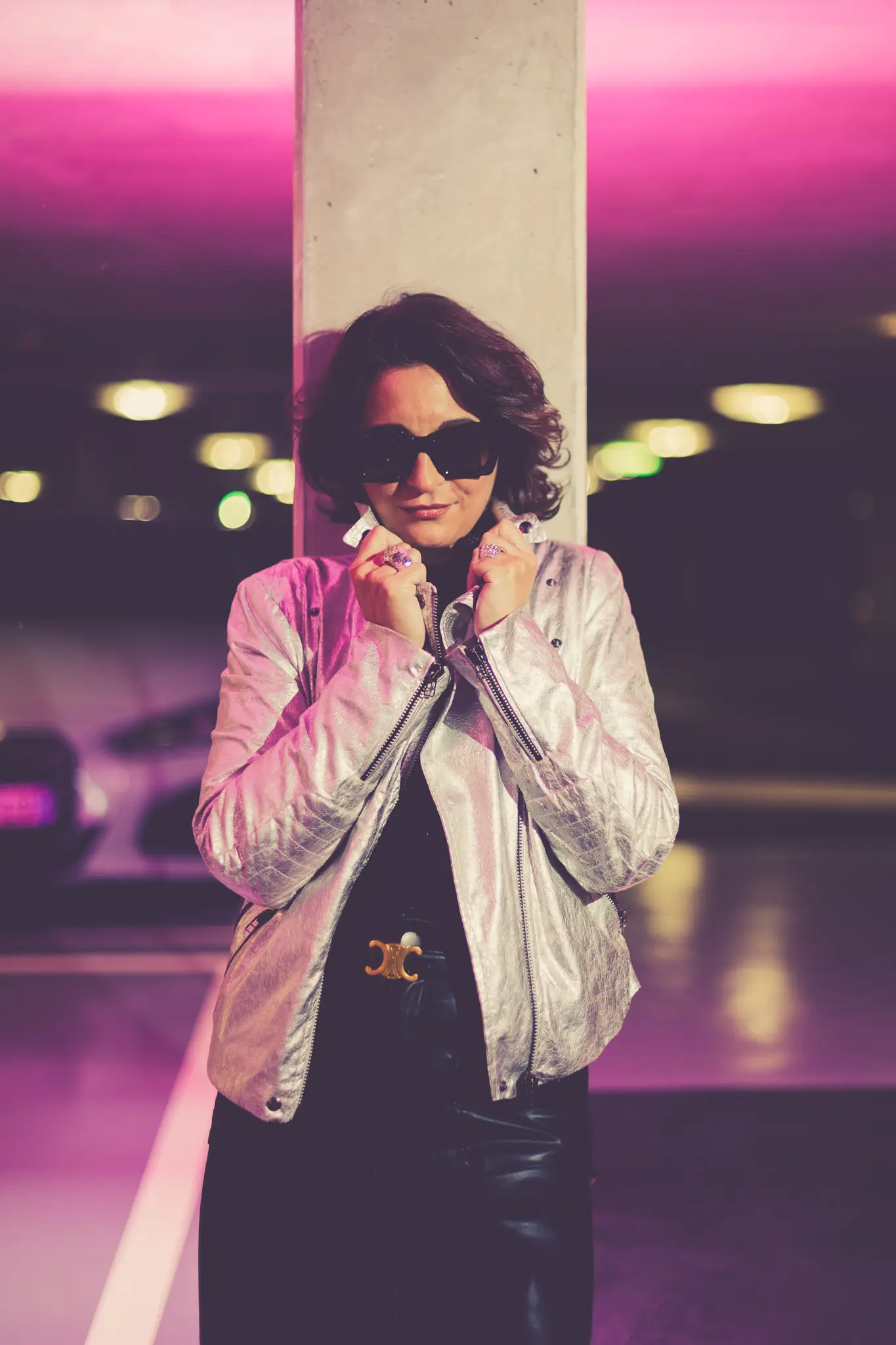 Silhouette rétro futuriste en veste métallisée argent posant sous des lumières néon dans un parking urbain.