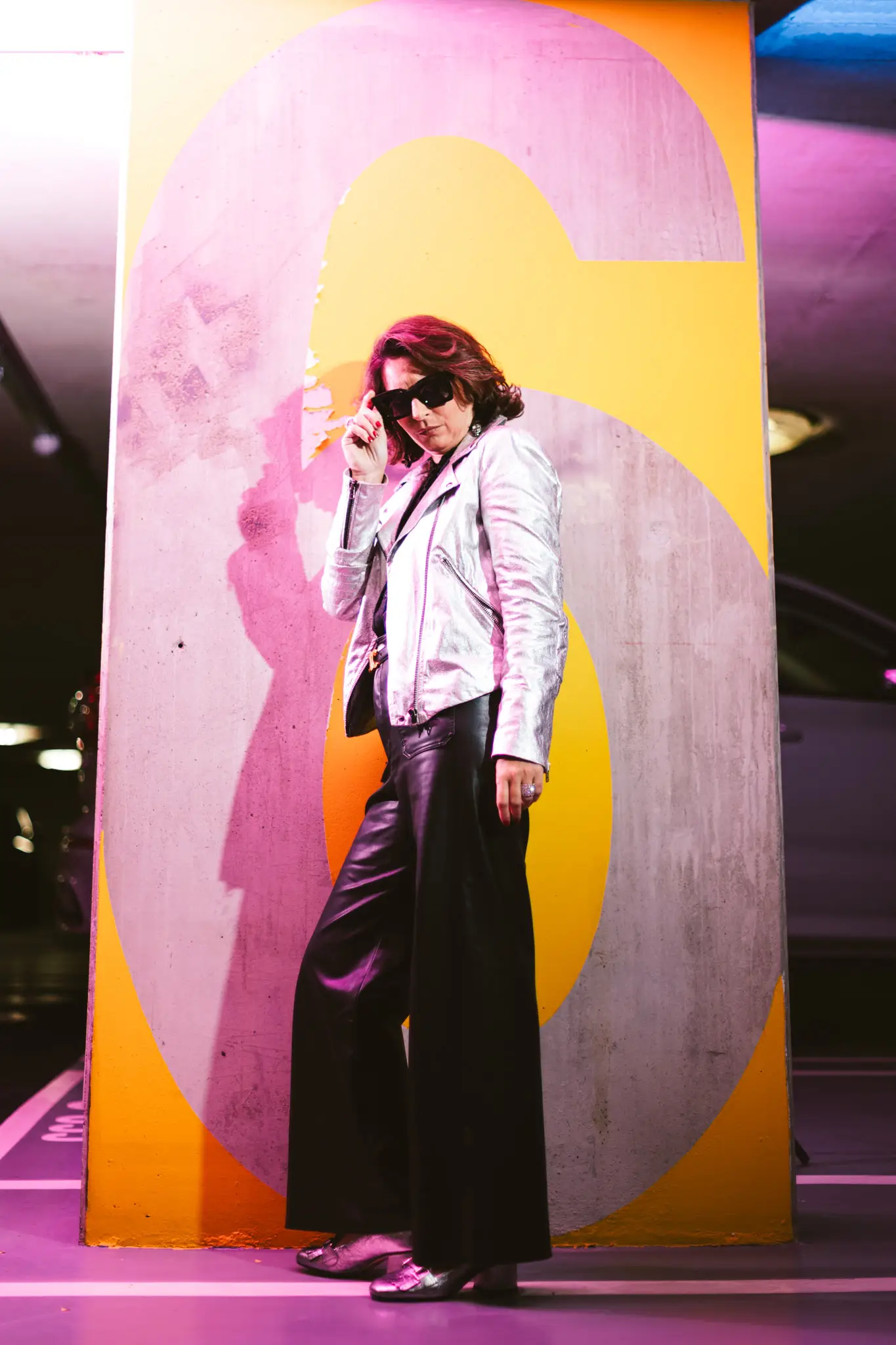 Silhouette rétro futuriste en veste métallisée argent et lunettes oversize posant sous des néons dans un parking urbain.