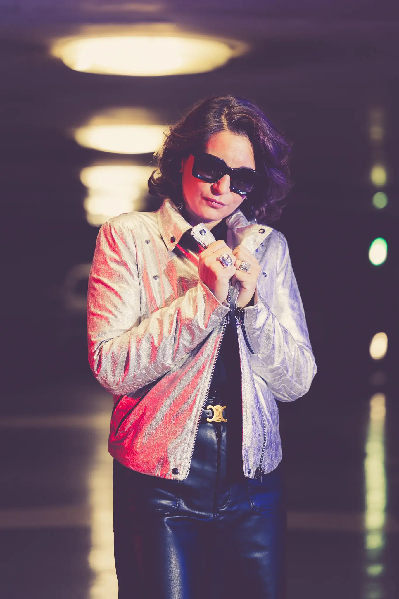 Silhouette rétro futuriste en veste métallisée argent et lunettes oversize dans un parking urbain.