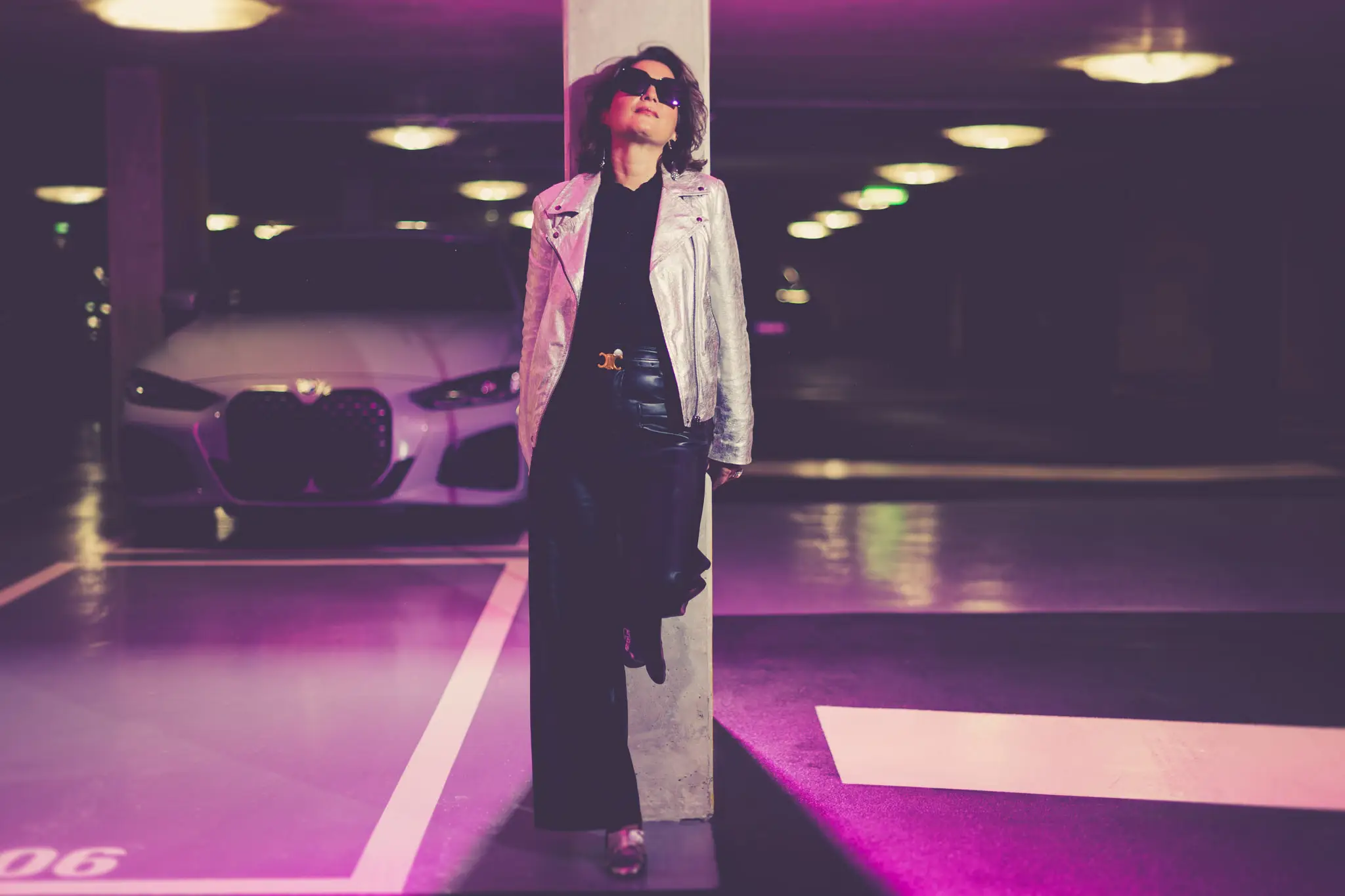 Silhouette mode rétro futuriste en veste métallisée posant sous lumières néon dans un parking, BMW i4 en arrière-plan.